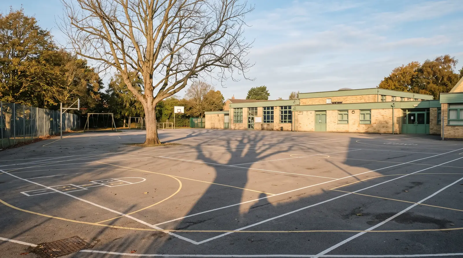 Cour d’école déserte au petit matin, lignes blanches et jaunes fraîchement tracées sur l’enrobé, longue ombre d’un arbre en travers de la cour